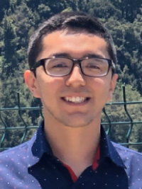 Carlos, sporting tussled black hair, standing outside wearing dark-rimmed glasses, and smiling